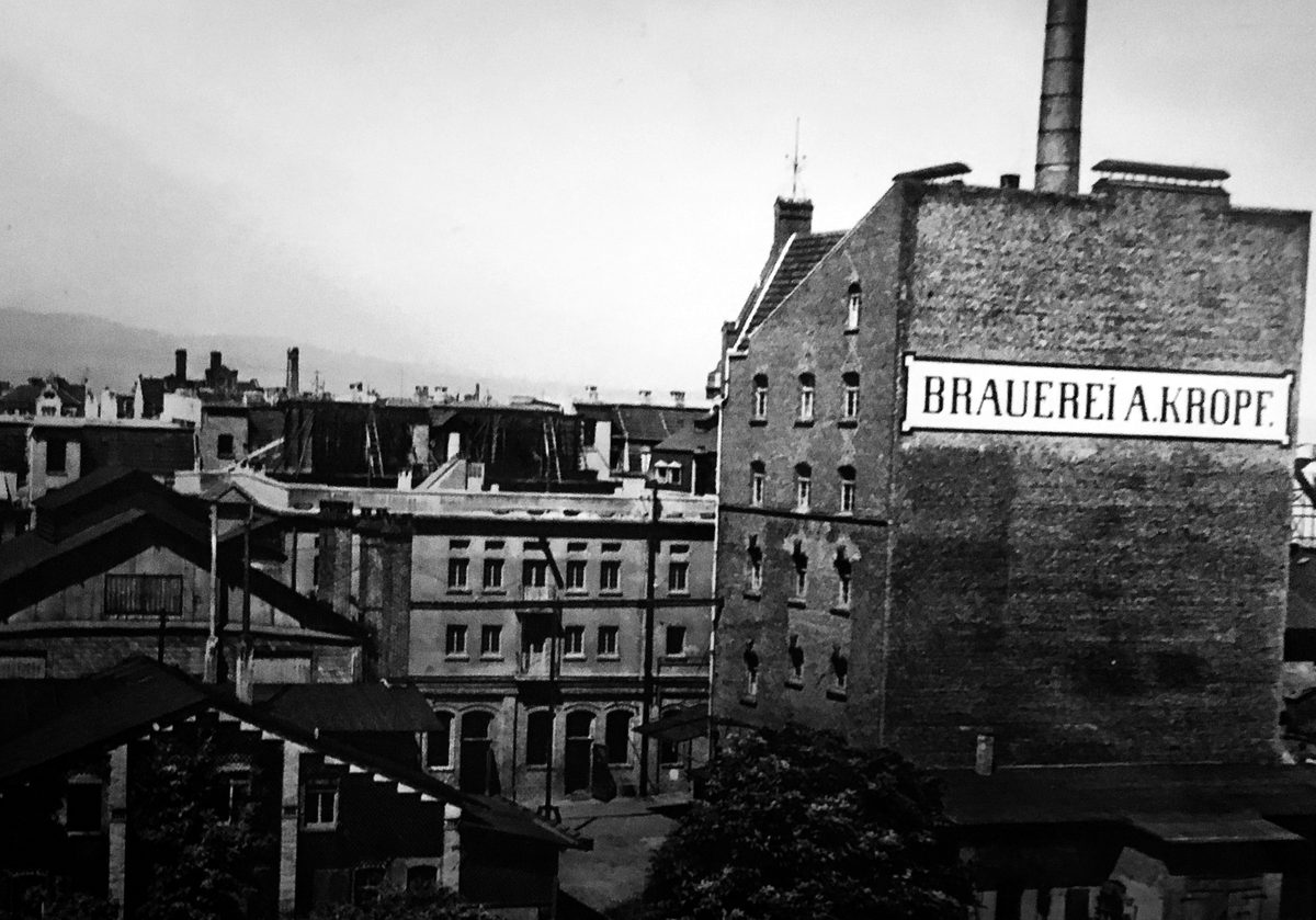 Historische Aufnahme: hoher Backsteinbau mit großem Schriftfeld „Brauerei A. Kropf“, dahinter ein Schornstein, links die Dächer der Umgebung.
